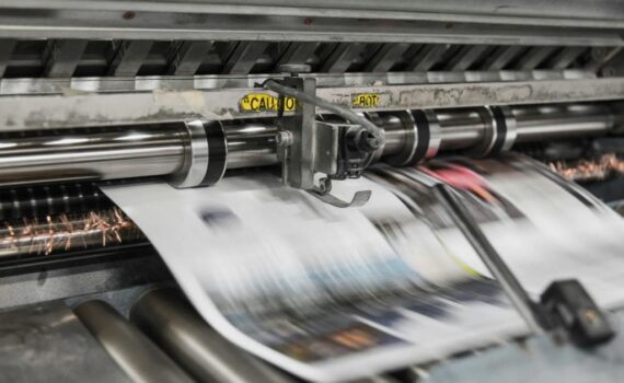 A close-up, high-speed industrial photograph of a professional printing press in motion. Large white sheets of paper with printed colorful graphics are shown blurring as they move rapidly through steel rollers and precision machinery. The setting is a clean, metallic industrial environment, emphasizing speed, technical accuracy, and high-volume production.