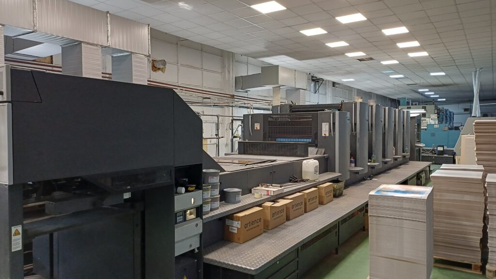 A wide-angle, high-tech shot of an 8-color sheetfed offset printing press operating in a clean, modern manufacturing facility, showing paper flowing from the feeder.