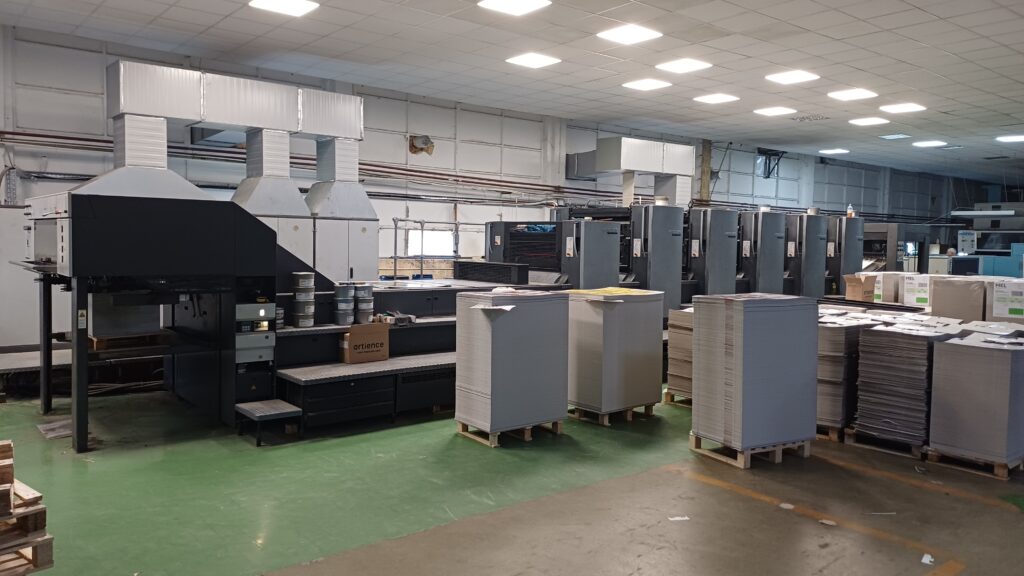 Large-scale industrial sheetfed offset printing press on a production floor with stacked pallets of printed substrates.