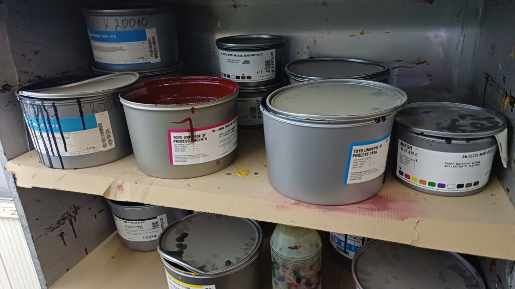 Open and closed cans of offset printing inks, including Toyo Process Magenta and Cyan, stored on a shelf in a commercial print shop environment.
