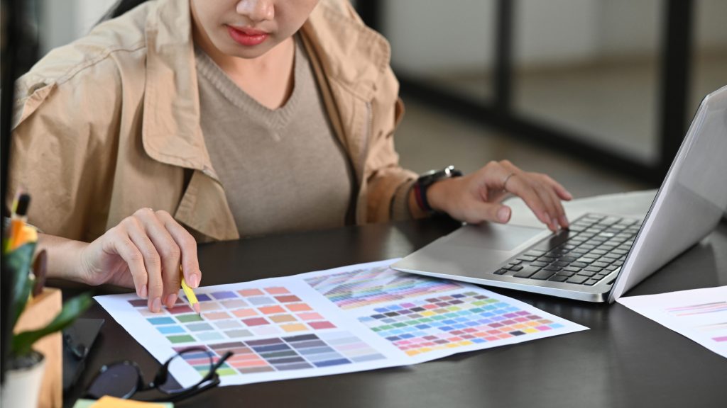 A prepress professional verifying color accuracy by comparing physical printed color swatches with digital source files on a laptop during the contract proofing phase.